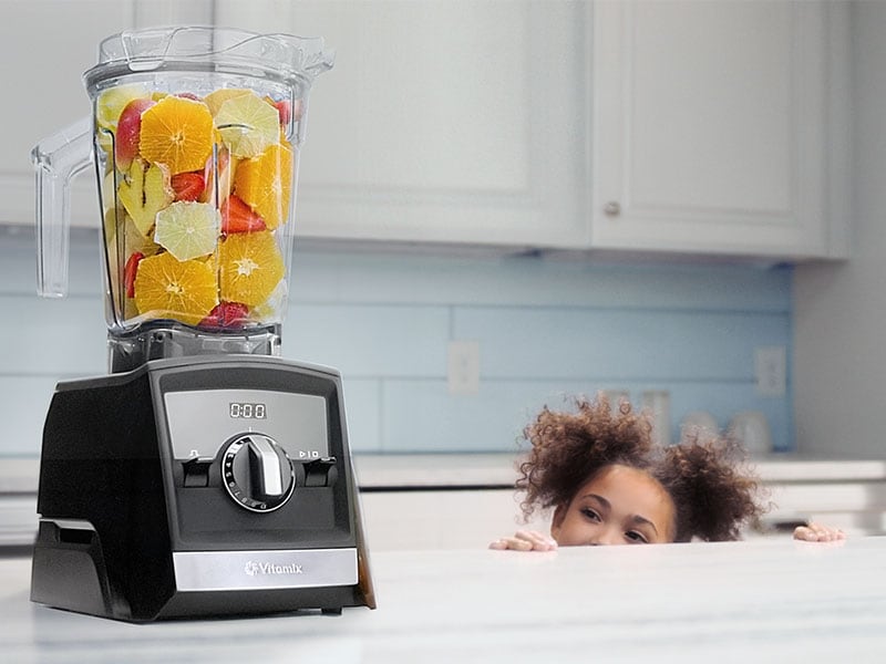 blender on counter with a smoothie build and a little girl looking up at it