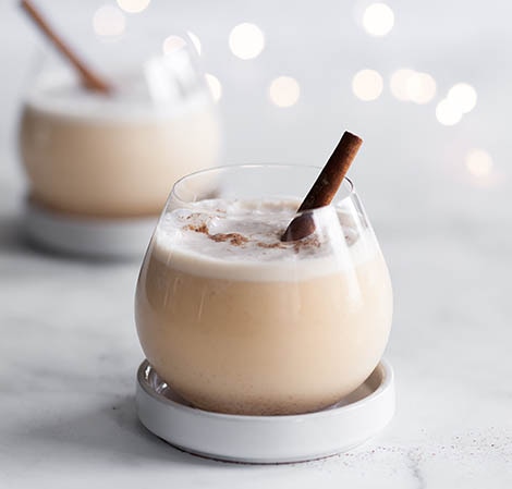 Eggnog in a glass with a cinnamon stick in it with twinkly lights in the background