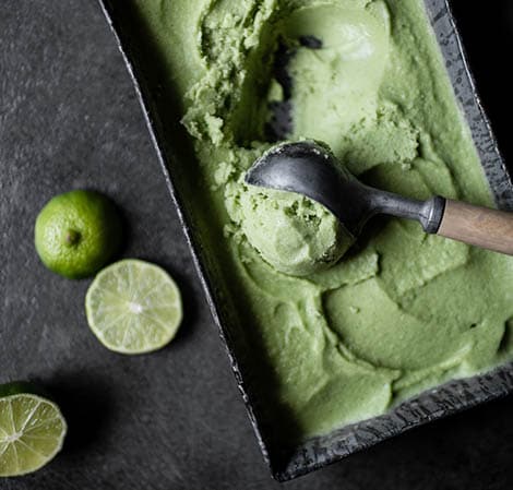 Key lime pie ice cream in a rectangular vessel being scooped out with some limes off to the side.