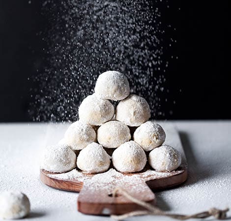 snowball cookies on a wooden tray with powdered sugar being sprinkled over them